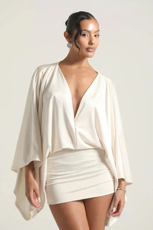 Woman wearing a white dress with a deep neckline and cape-like sleeves against a plain background