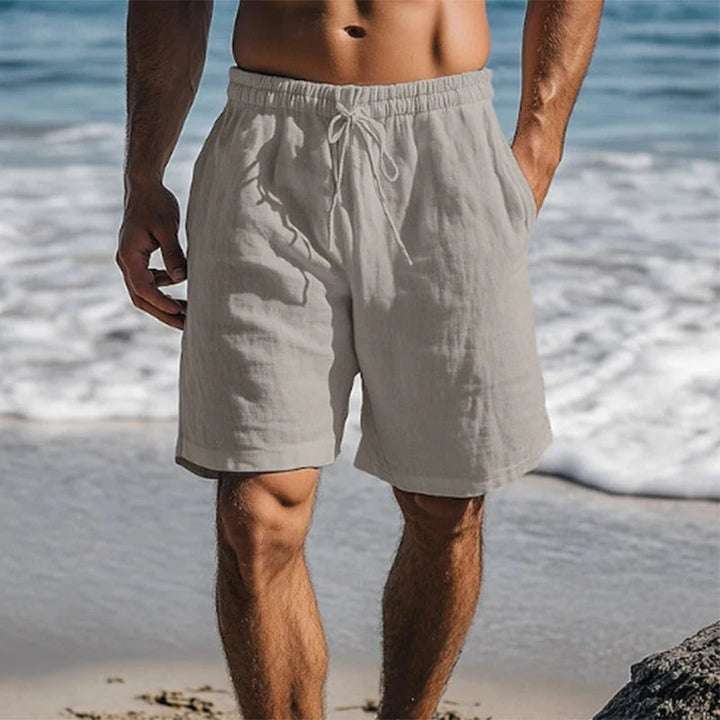Light gray drawstring cotton beach shorts shown on a man at the shoreline