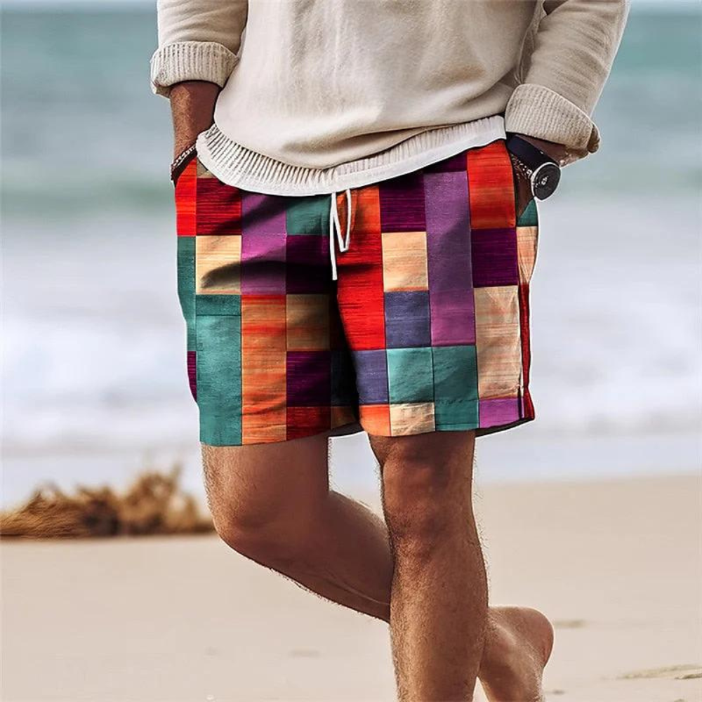 Men's swimsuit board shorts with pockets in red purple teal patchwork print, beach front view