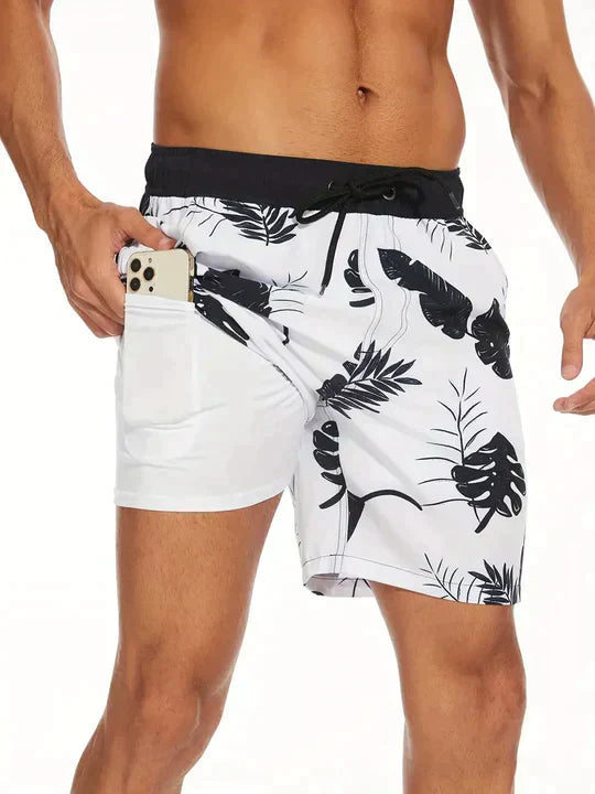 White tropical leaf men's swim shorts with black waistband and side pocket holding phone
