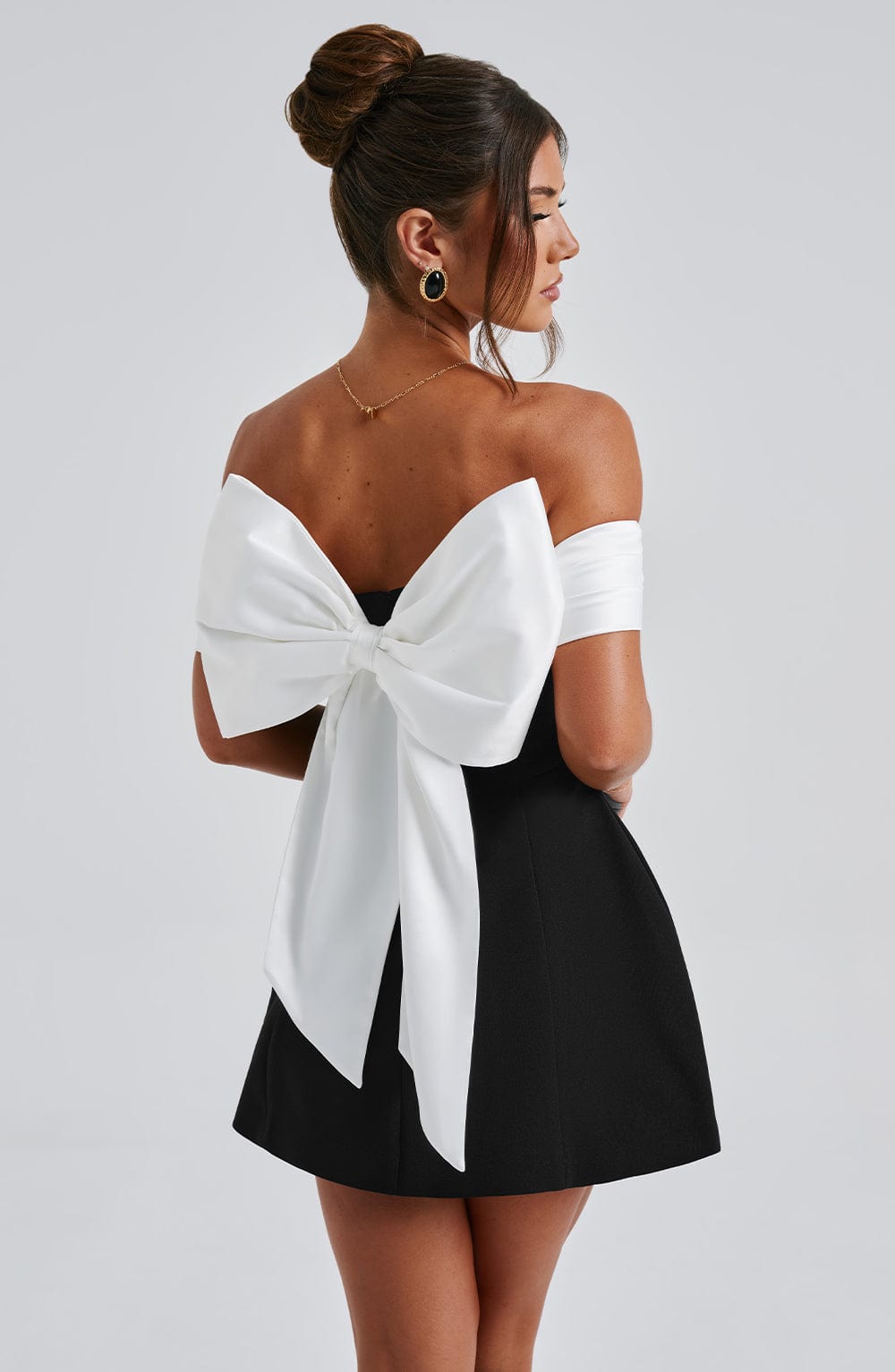 Back angled view of black off shoulder mini dress with large white bow and open back detail