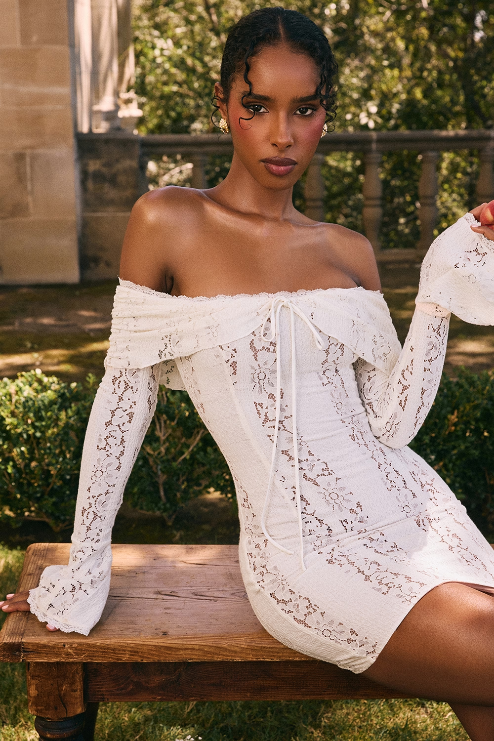 Model seated in off shoulder lace mini dress with long sleeves and floral lace detail